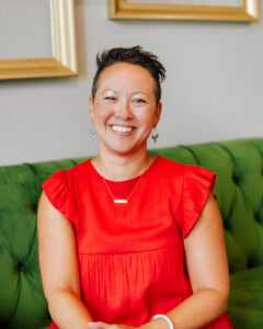 Jackie Murphy, social media coach, wears a bright red dress while sitting on the green coach at Haven Collective and smiling.