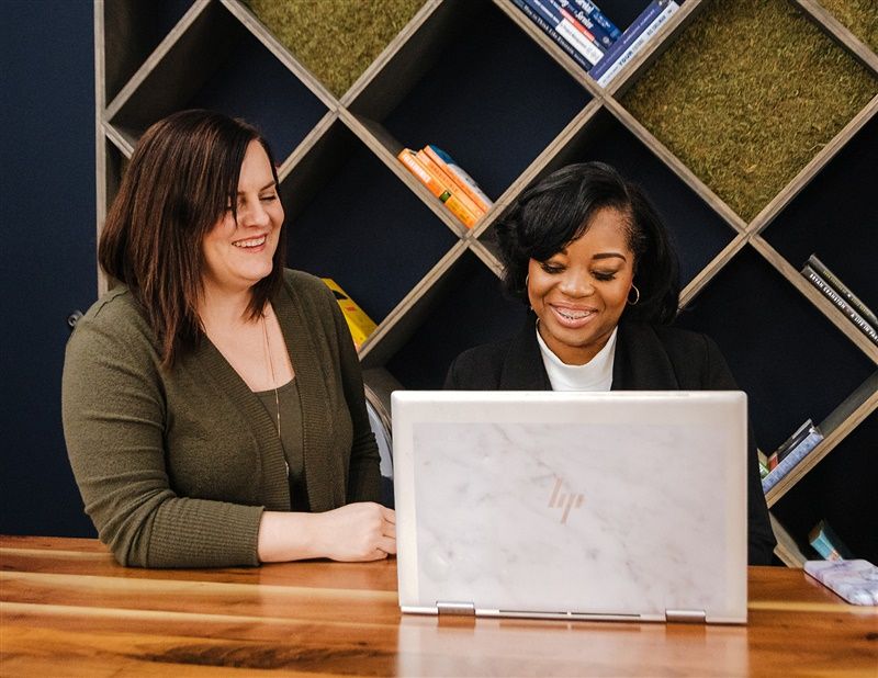 Two women sit at a desk behind a computer, smiling