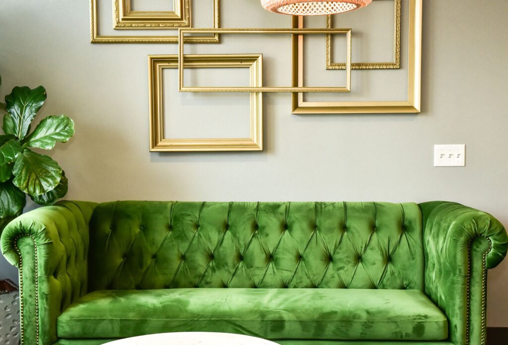 Green velvet sofa in front of grey wall that has gold picture frames hanging on it